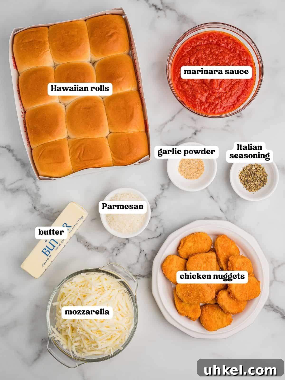 Chicken Parmesan Sliders Recipe: Crispy Mini Sandwiches 4 Labeled ingredients for chicken parm sliders on a marble surface for chicken parmesan sliders, including a package of Hawaiian rolls, a bowl of marinara sauce, a stick of butter, a bowl of shredded mozzarella, a bowl of grated Parmesan, a plate of chicken nuggets, and small bowls of garlic powder and Italian seasoning.