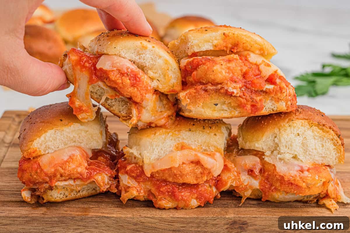 Chicken Parmesan Sliders Recipe: Crispy Mini Sandwiches 7 Chicken parmesan sliders are stacked on a wooden board. Each sandwich contains a breaded chicken nugget, marinara sauce, and melted cheese, with slightly toasted, herb-sprinkled buns. A hand is lifting one from the stack.