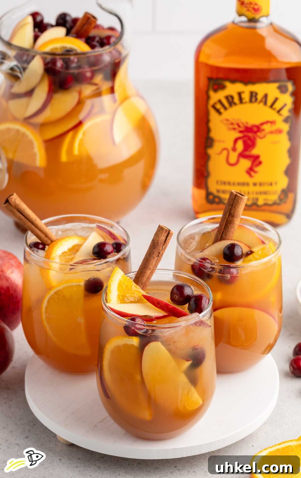 5-Minute Fireball Sangria Recipe for Quick Party Drinks 8 Three glasses of Fireball apple cider sangria with fruit slices and cinnamon sticks next to a large pitcher and bottle of Fireball.