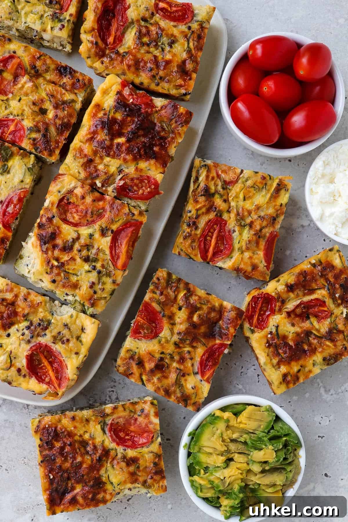 Some cut up zucchini slice squares on a plate and some on the table. Small dishes of cottage cheese, mashed avocado and cherry tomatoes for decoration.