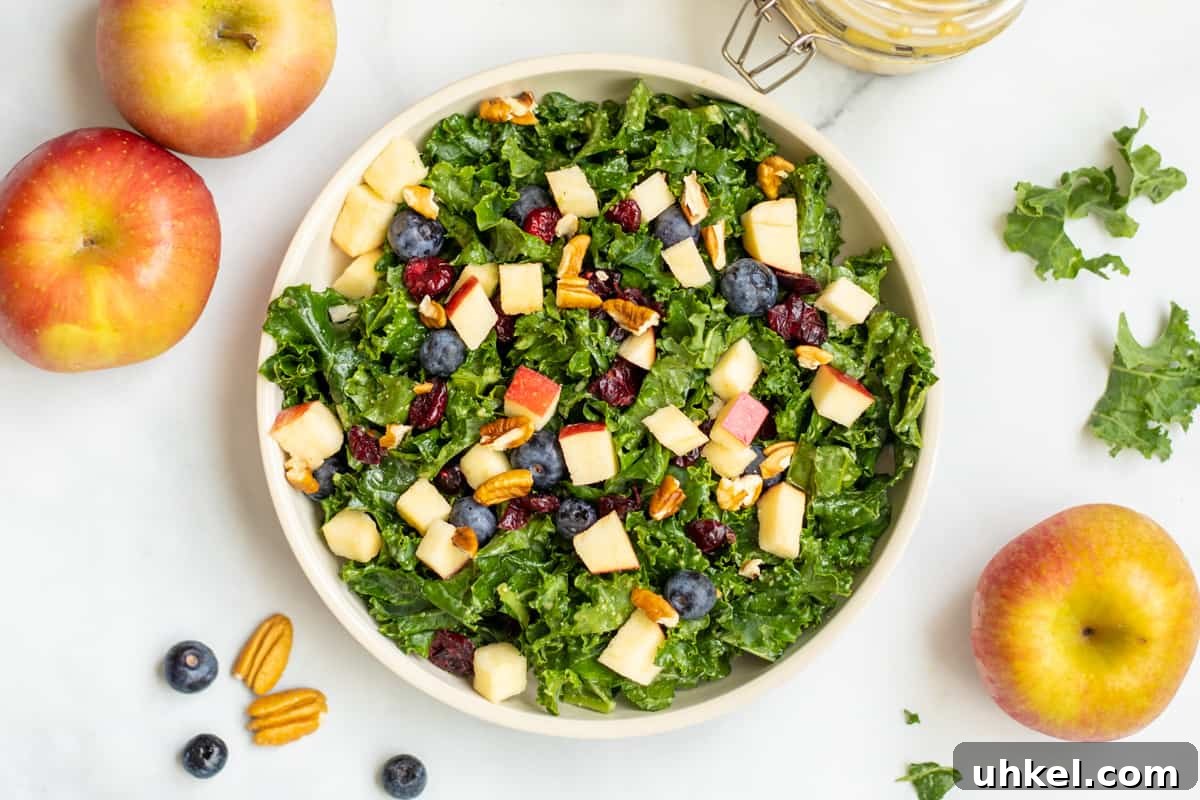 Massaged Kale Salad Kale salad with apples, berries and nuts in bowl with pecans and apples in background.