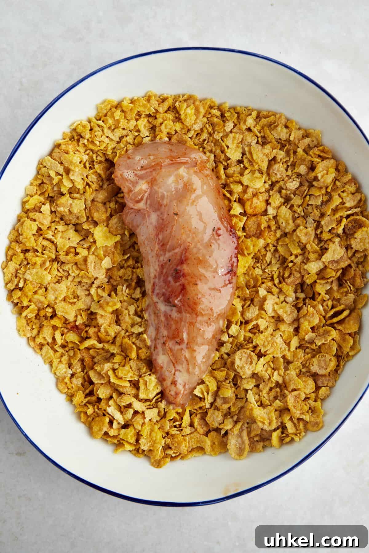 Raw chicken coated with eggs wash in a bowl of crushed cornflakes.