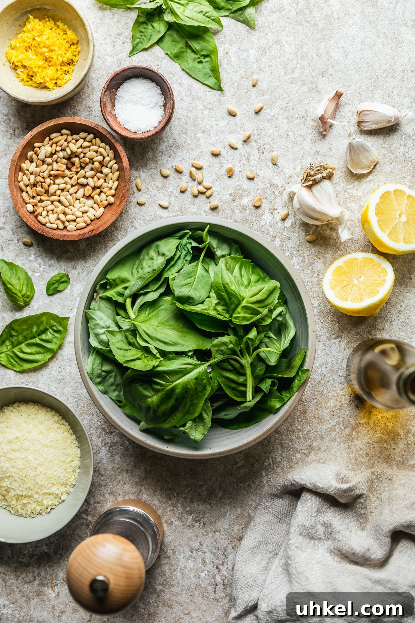 Lemon Basil Pesto Recipe — Bright Sauce for Pasta & Sandwiches 3 A grey bowl of basil on a beige counter next to halved lemons, a jar of olive oil, a beige linen, a grey bowl of Parmesan, a white bowl of lemon zest, and a wood bowl of pine nuts.