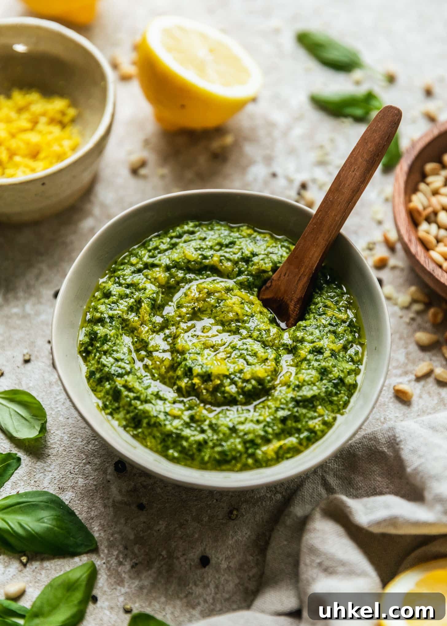 Lemon Basil Pesto Recipe — Bright Sauce for Pasta & Sandwiches 6 A grey bowl of lemon basil pesto on a beige counter next to halved lemons, a wood bowl of pine nuts, a white bowl of lemon zest, and basil leaves.