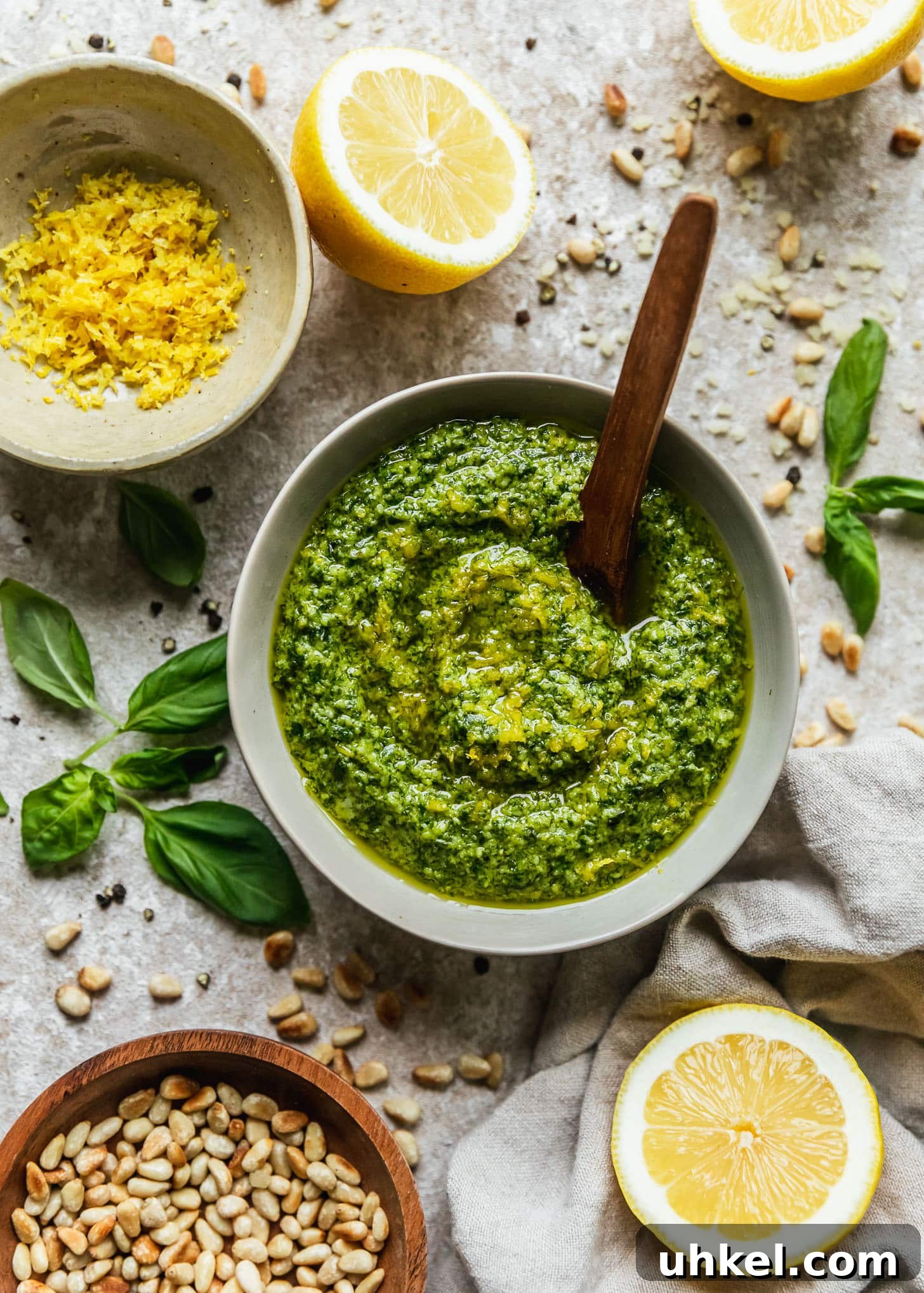 Lemon Basil Pesto Recipe — Bright Sauce for Pasta & Sandwiches 7 A grey bowl of lemon pesto with a wooden spoon on a beige counter next to lemon halves, basil leaves, a white bowl of lemon zest, a wood bowl of pine nuts, and a beige linen.
