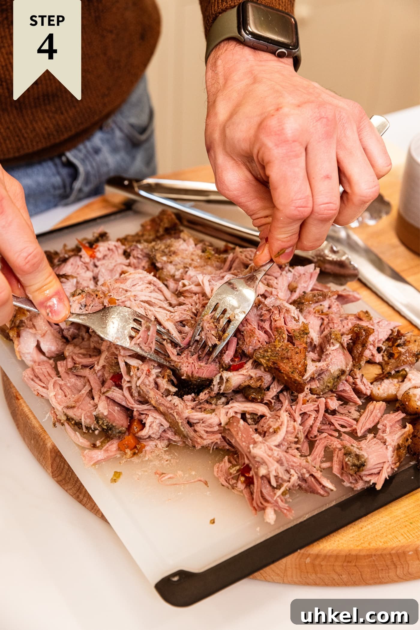 Slow-Cooked Italian Roast Pork Sandwich Recipe 11 Hands holding two forks shredding pork shoulder into individual pieces on plastic cutting board with lots of meat and tongs sitting to the side.