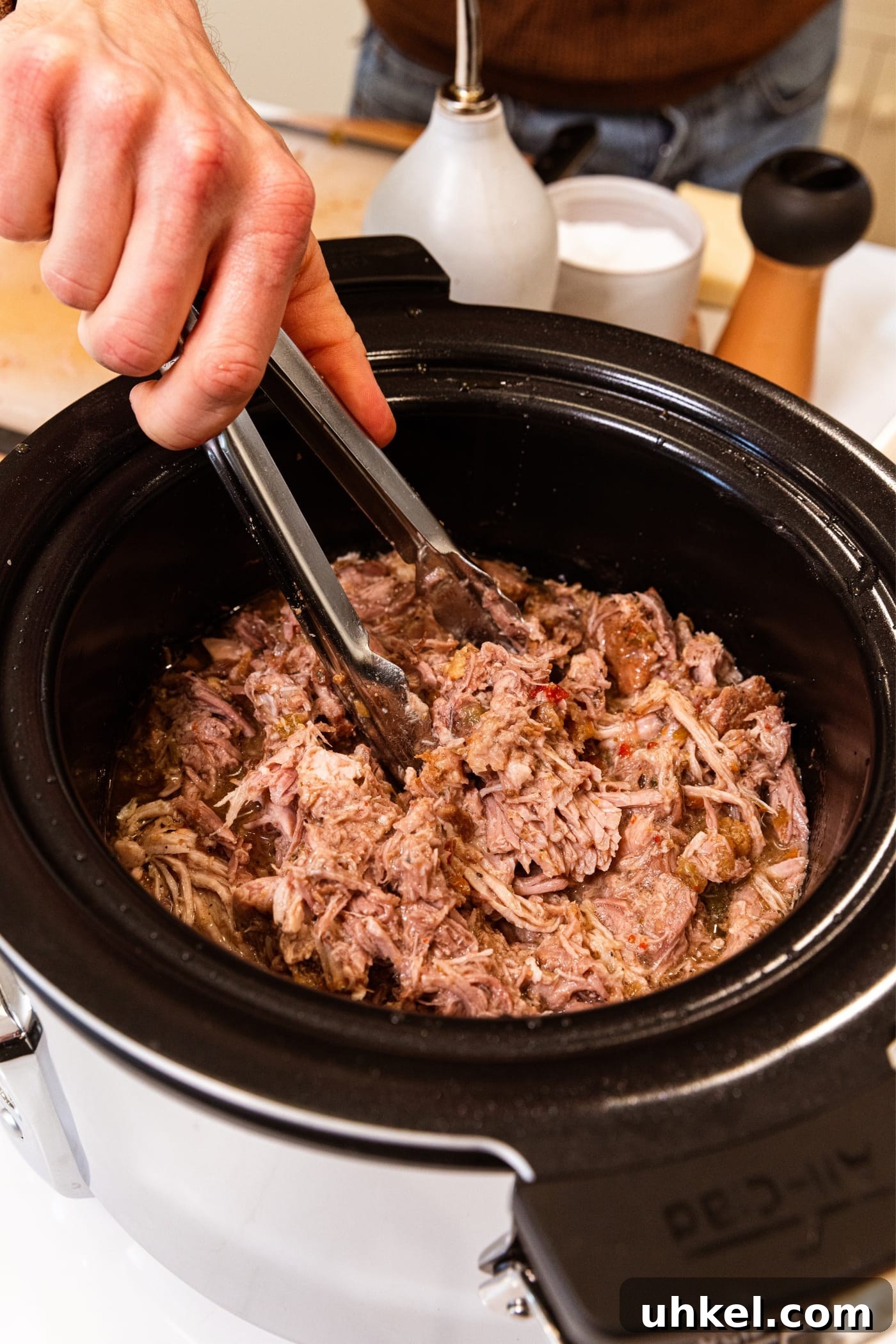 Slow-Cooked Italian Roast Pork Sandwich Recipe 12 Hand holding tongs stirring shredded pork in slow cooker bowl with liquid pushing up on the sides.