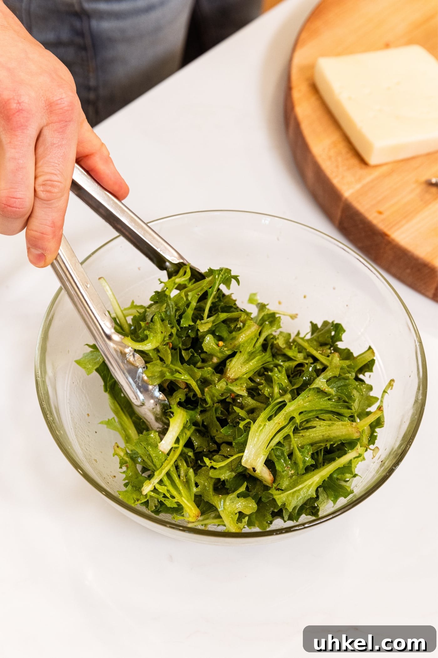Slow-Cooked Italian Roast Pork Sandwich Recipe 14 Hand using tongs to mix greens into Italian dressing in medium-sized glass bowl on white countertop with wood cutting board in background.
