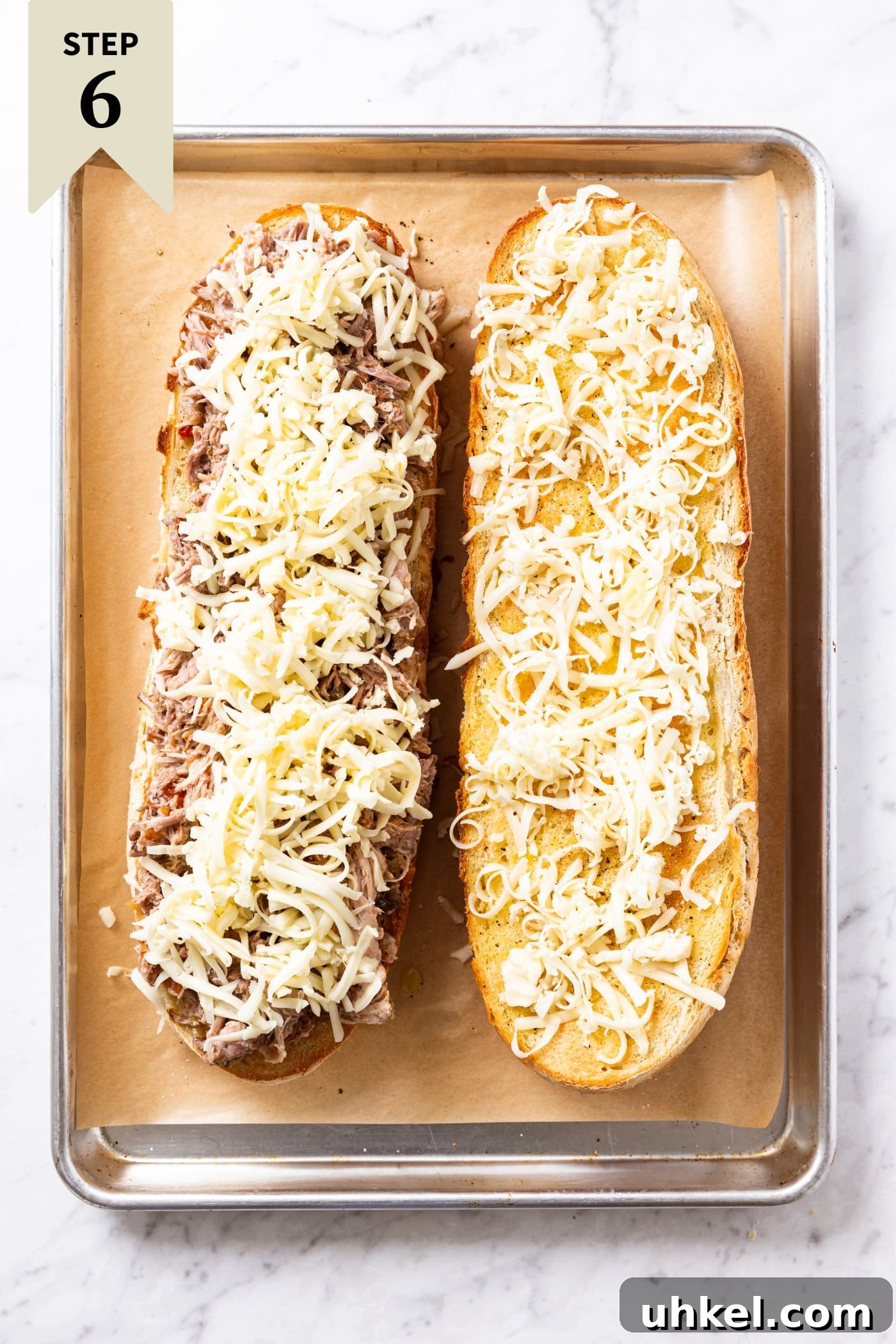 Slow-Cooked Italian Roast Pork Sandwich Recipe 15 White marble surface with parchment-lined sheet pan filled with two halves of French bread sitting open faced with one piled with meat and the other piled with shredded cheese right before going into the oven.