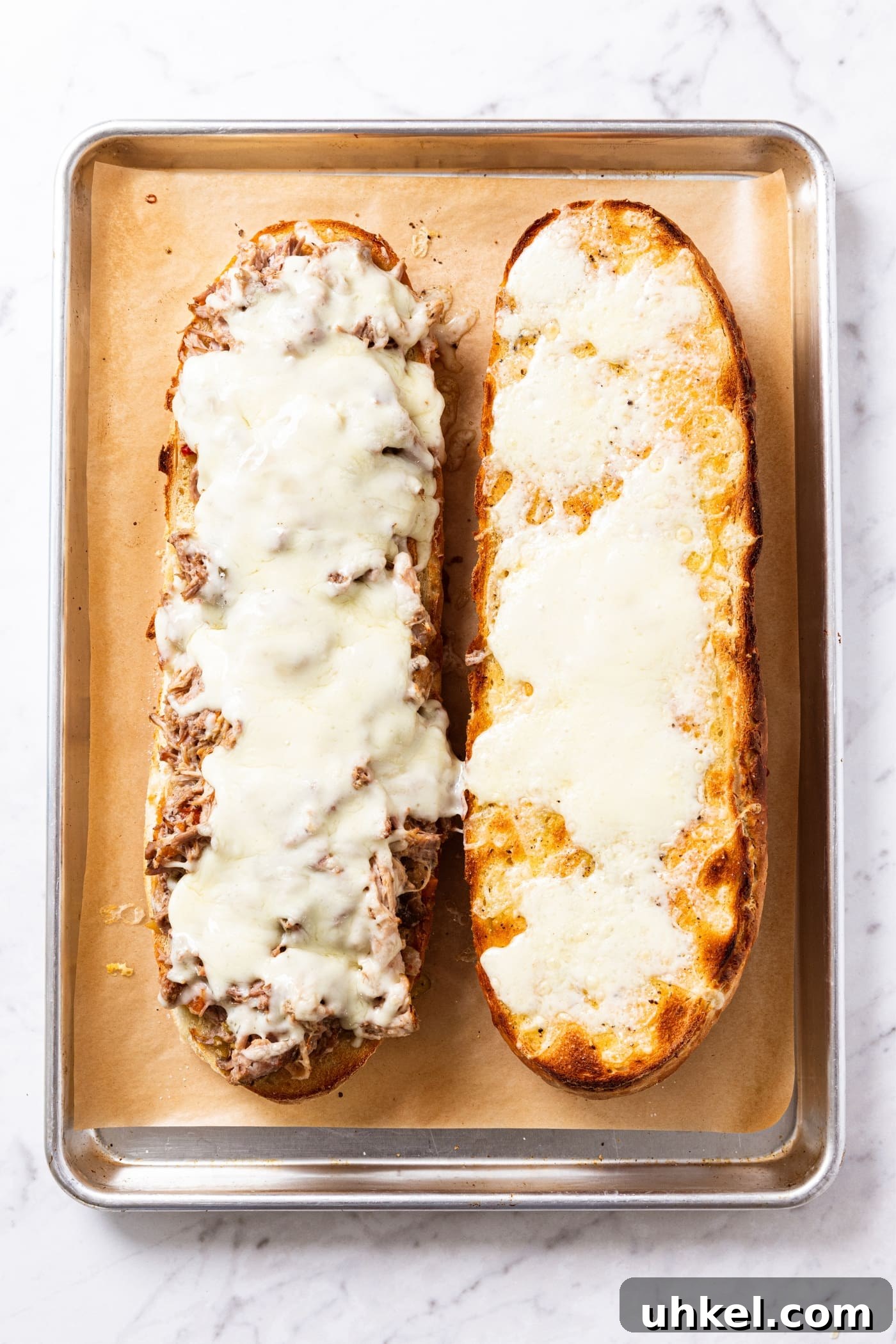 Slow-Cooked Italian Roast Pork Sandwich Recipe 16 White marble surface with parchment-lined baking sheet with two halves of French bread after coming out of the oven after being toasted with melted cheese all over the top.