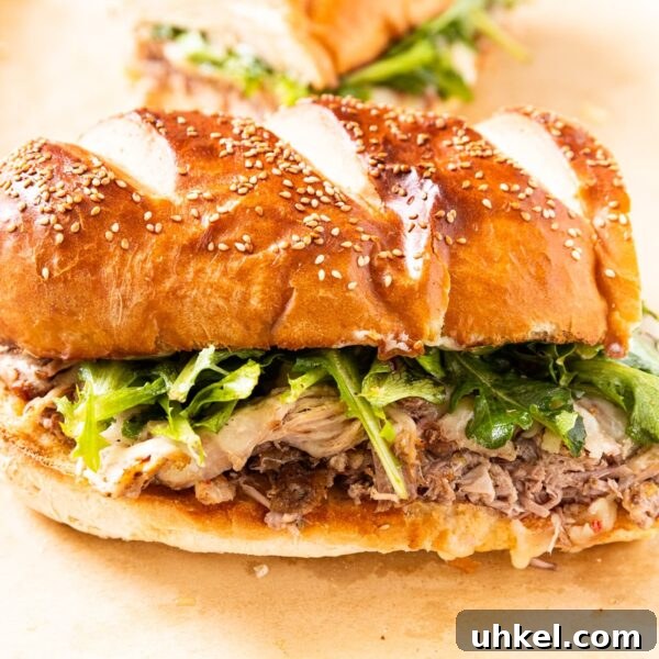 Slow-Cooked Italian Roast Pork Sandwich Recipe 18 Half of a roasted pork sandwich topped with greens and cheese on French bread sitting on a piece of brown-colored parchment paper on white surface.