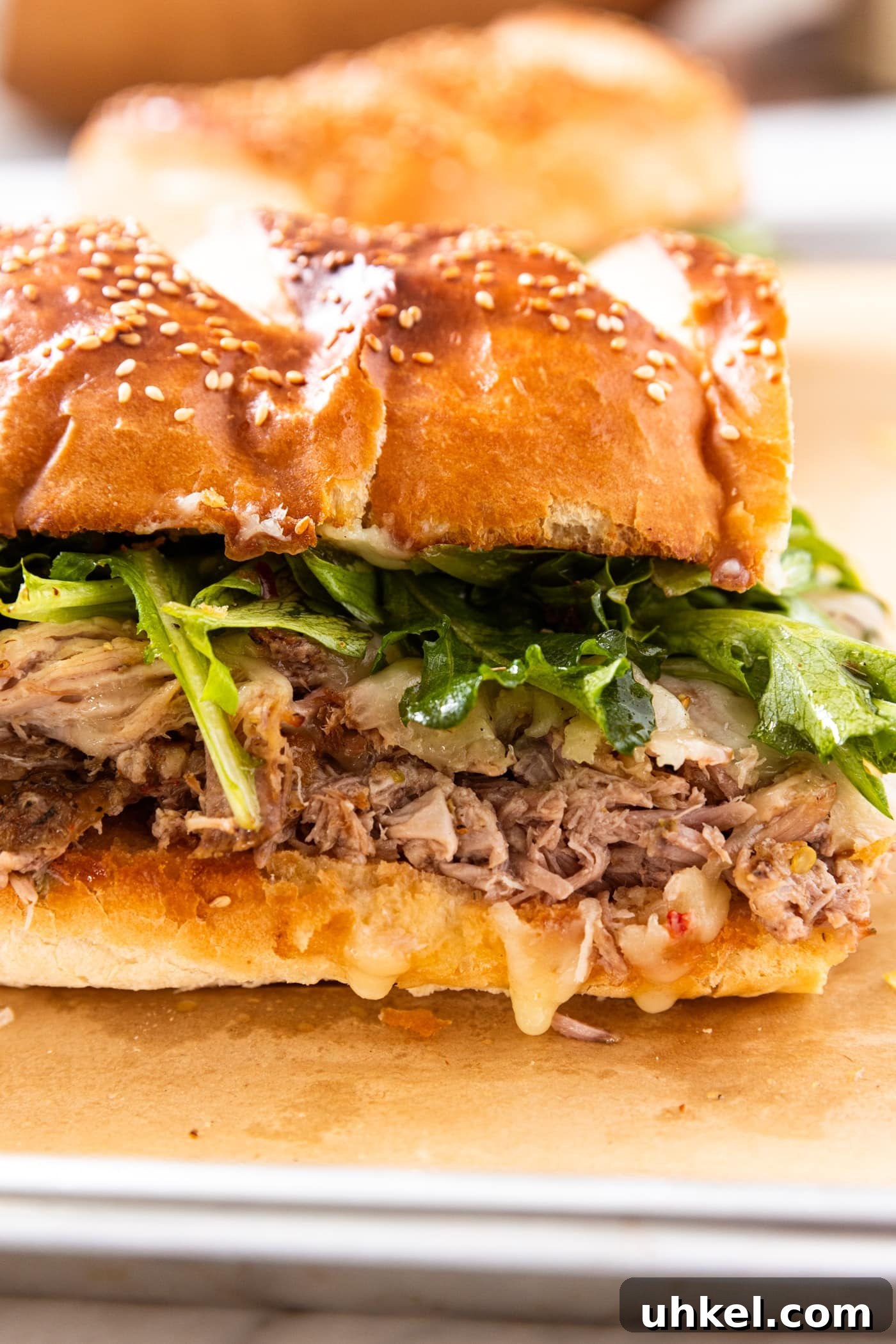 Slow-Cooked Italian Roast Pork Sandwich Recipe 3 Italian roasted pork sandwich full of pulled pork and topped with greens and cheese sitting on a parchment-lined baking sheet after being toasted in the oven.