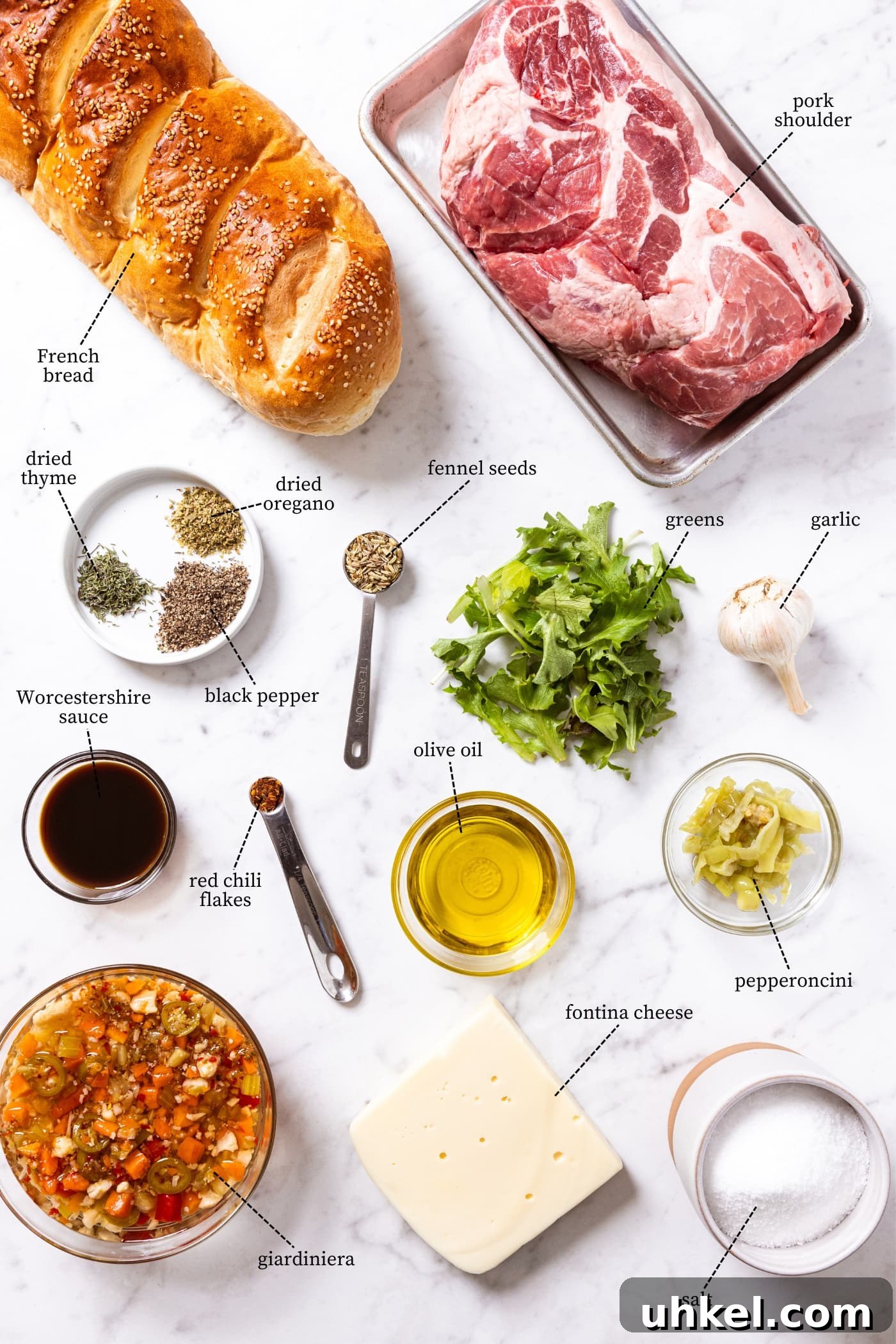 Slow-Cooked Italian Roast Pork Sandwich Recipe 4 White marble surface filled with all ingredients needed to make roasted pork sandwich including French bread, pork shoulder, spices, cheese, salt, Worcestershire sauce, greens, and more.