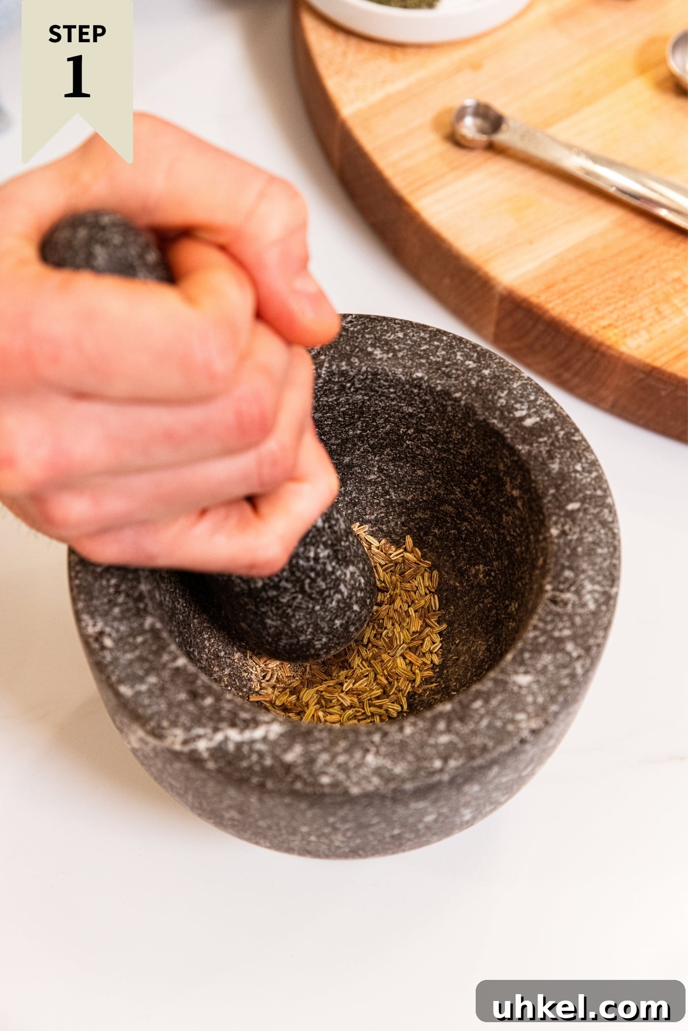 Slow-Cooked Italian Roast Pork Sandwich Recipe 5 Hand holding pestle using it to grind fennel seed in a gray mortar on white countertop with wood cutting board in background.