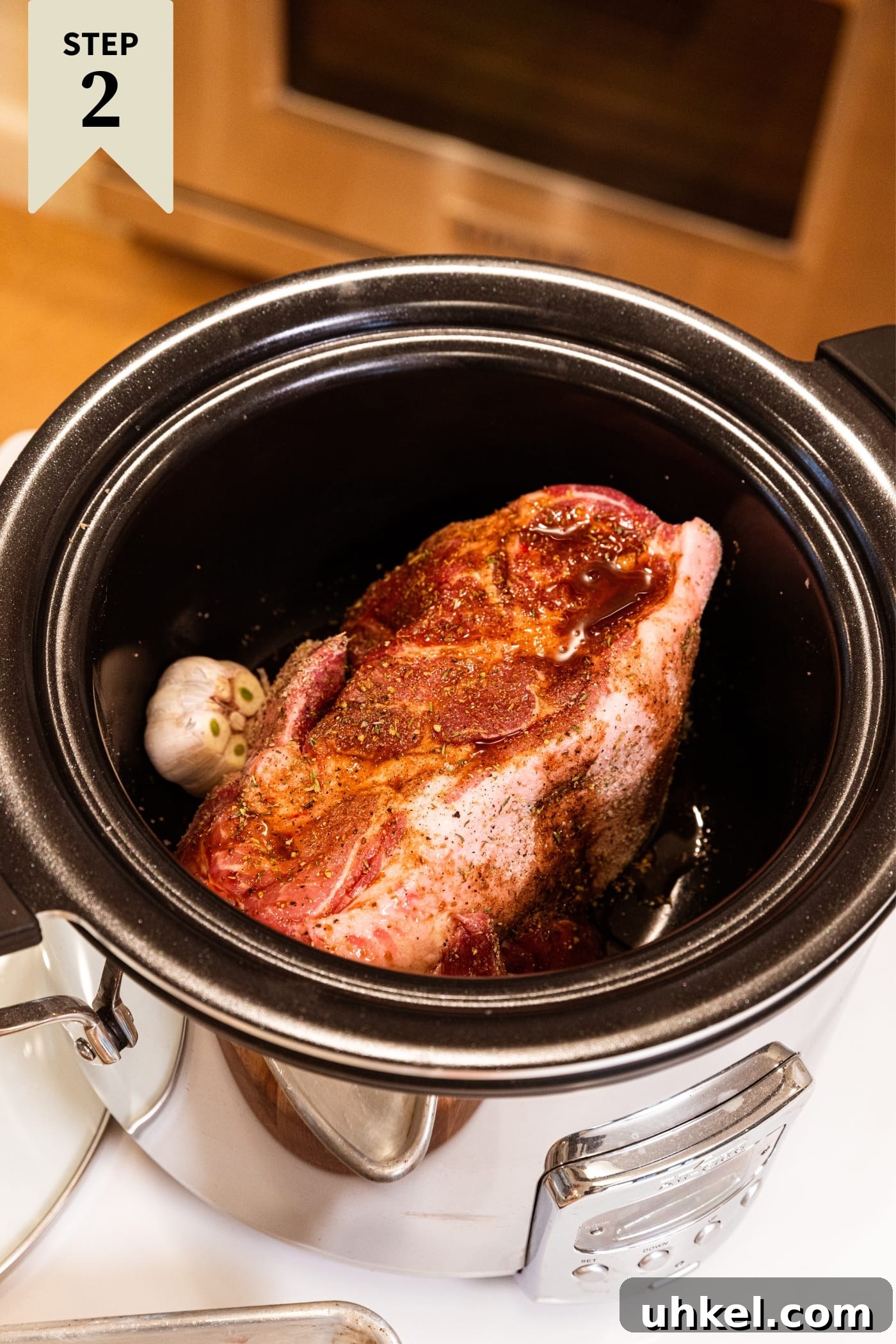 Slow-Cooked Italian Roast Pork Sandwich Recipe 7 Slow cooker sitting on white countertop with raw piece of pork should sitting in bowl with garlic head on the side and Worcestershire sauce poured all over the top with stove in background.