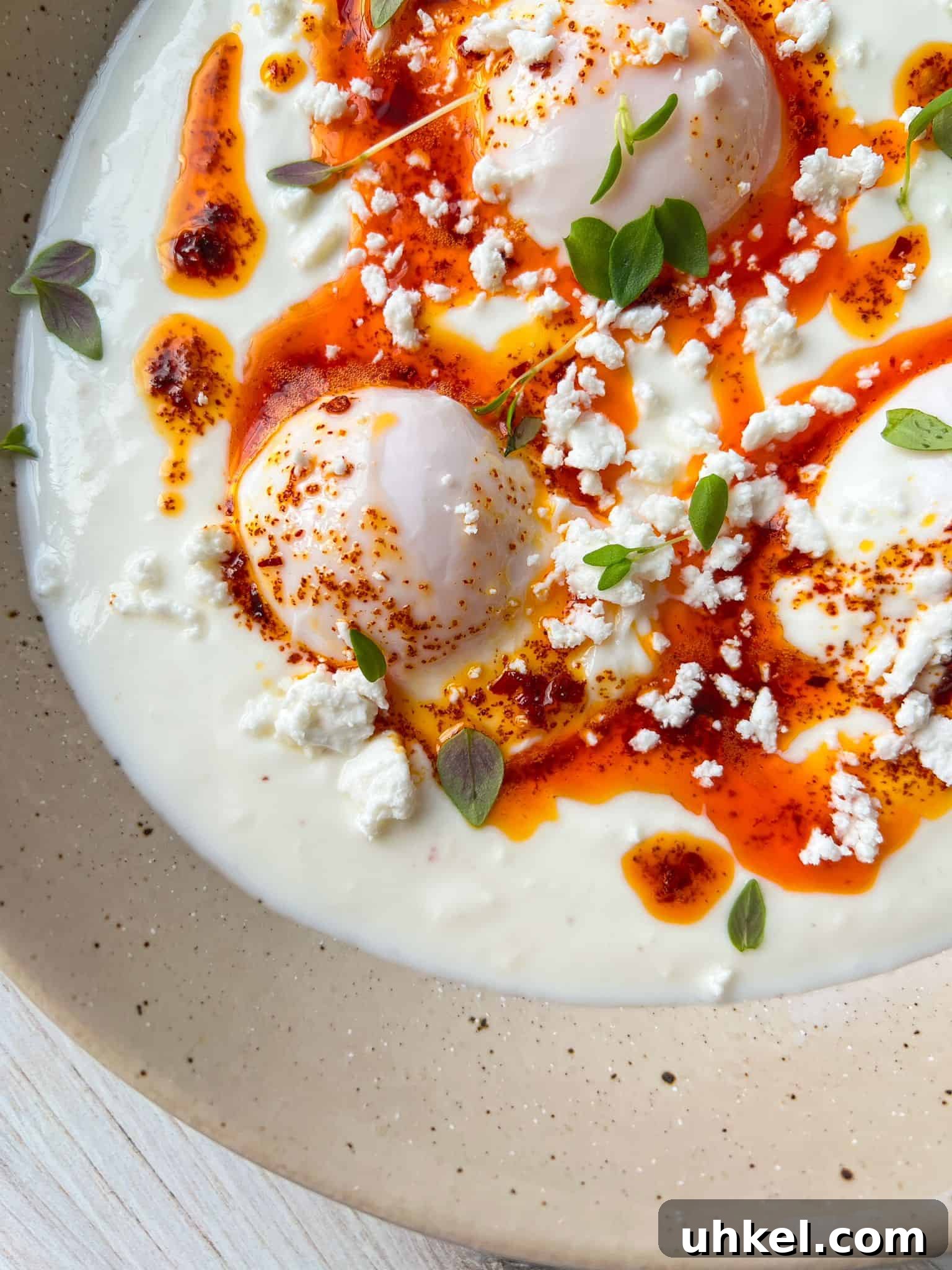 A close-up of poached eggs on a bed of creamy yogurt, drizzled with spicy red chili oil and garnished with fresh herbs and crumbled cheese, served on a speckled beige plate.