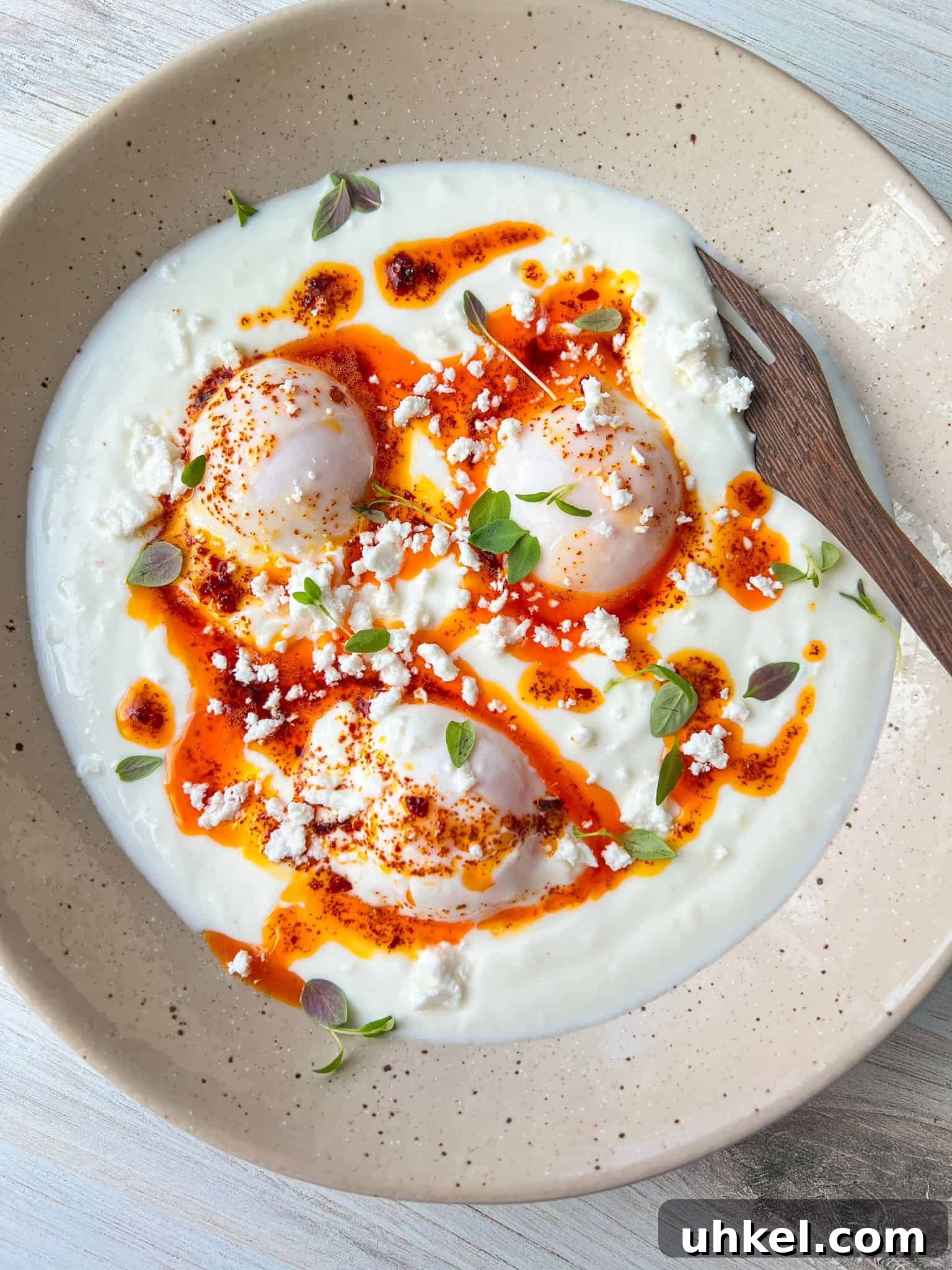 A dish featuring poached eggs on a bed of yogurt, drizzled with spicy red oil, garnished with crumbled feta cheese and fresh herbs. A wooden spoon rests on the beige plate.