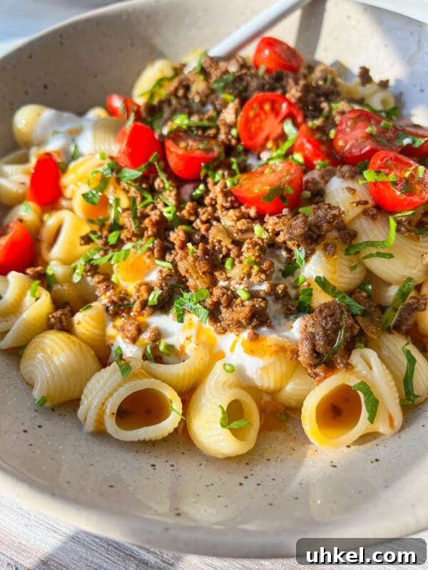 A bowl of pasta shells topped with ground meat, sliced cherry tomatoes, yogurt, and fresh herbs.