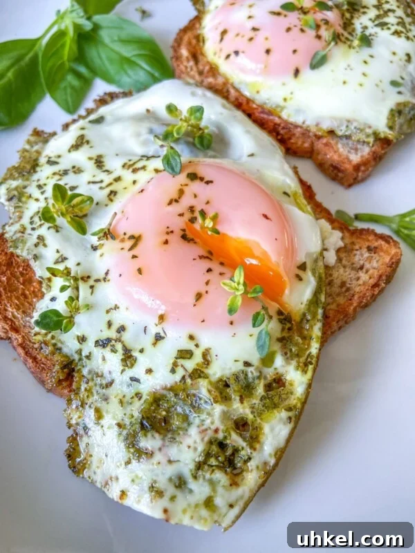 Two slices of toast topped with sunny-side-up eggs, green pesto, and sprinkled herbs.