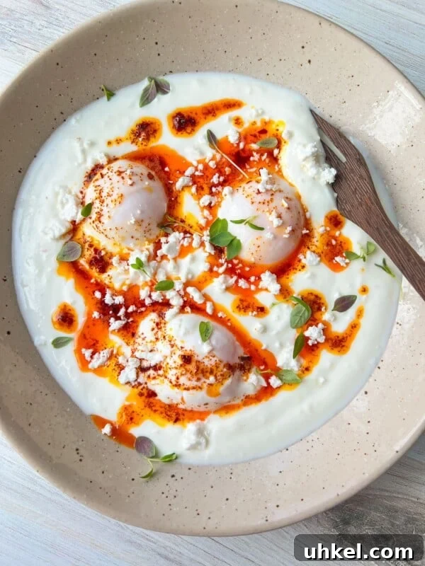 A dish featuring poached eggs on a bed of yogurt, drizzled with spicy red oil, garnished with crumbled feta cheese and fresh herbs.