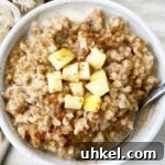 overhead shot of a slow cooker apple cinnamon oatmeal in a white bowl with diced apple on top & milk with a silver spoon