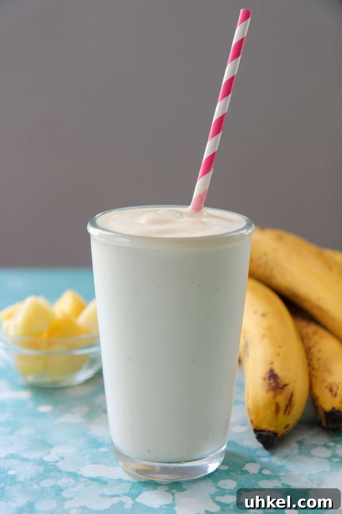 Creamy Pineapple Banana Smoothie Recipe for Tropical Energy 4 pineapple smoothie in a tall glass with a pink and white straw and bananas and pineapples in the background.