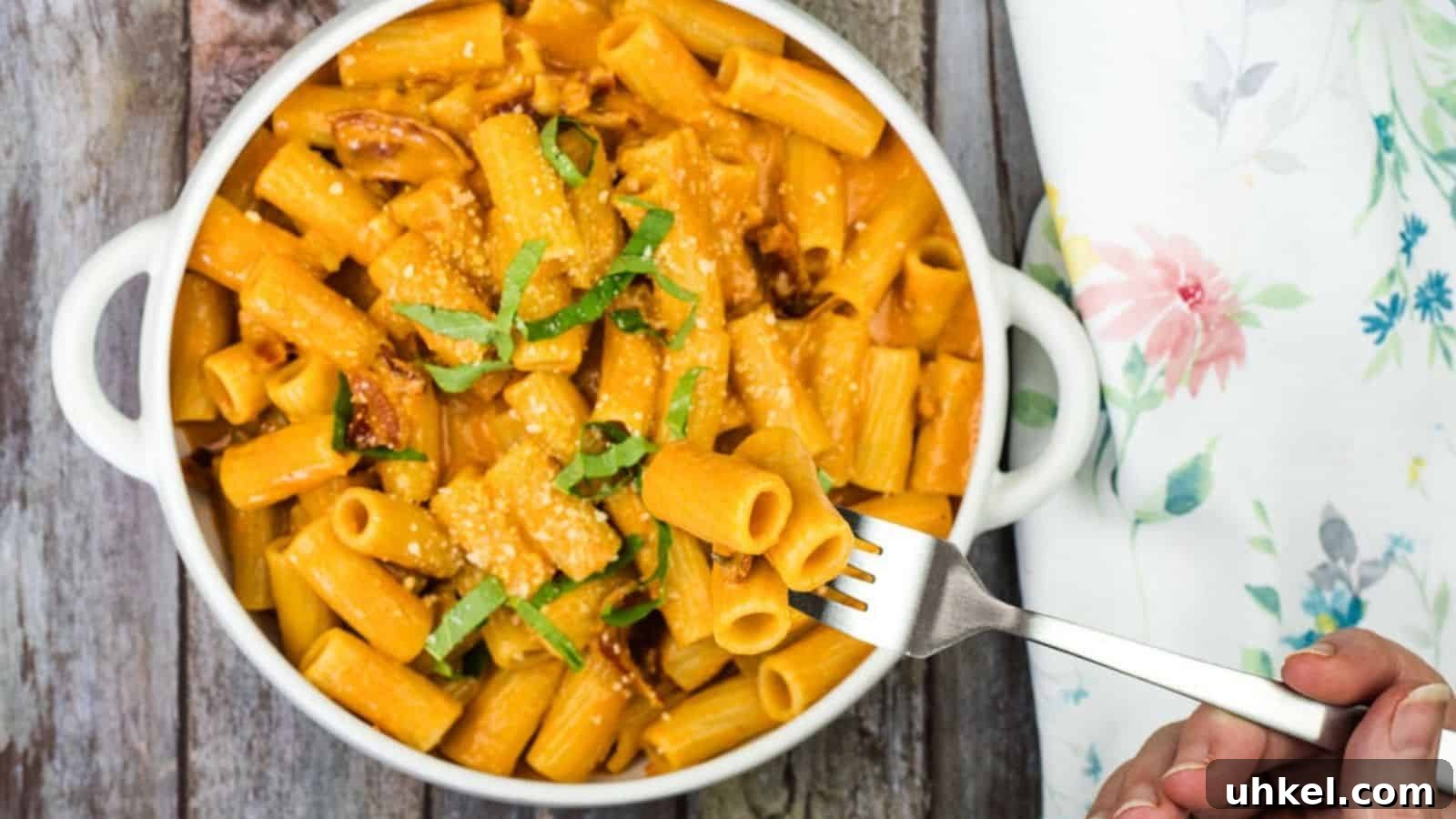 19 Budget-Friendly Dinners That Cut Your Grocery Bill (Delicious!) 12 A white bowl of rigatoni pasta with creamy sauce, garnished with cheese and green herbs, is placed on a wooden surface. A hand is holding a fork with a piece of pasta.