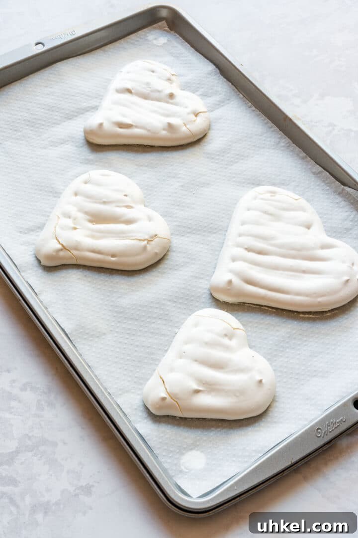 Heart-Shaped Mini Pavlovas: Valentine Dessert Recipe Ideas 9 Baked meringue hearts cooling on a parchment-lined baking sheet.