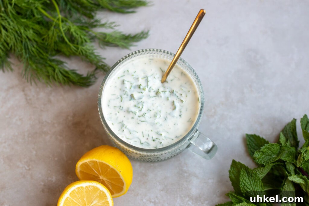 Vegan Creamy Yogurt and Herb Dressing Recipe 4 top view of a glass dish of vegan herb salad dressing made with vegan yogurt, dill and mint, lemon juice and maple syrup sits on a gray surface surrounded by fresh dill and mint leaves and a cut open lemon.