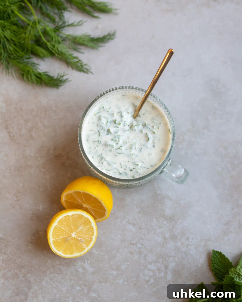 Vegan Creamy Yogurt and Herb Dressing Recipe 6 small bowl of yogurt and herbs dressing sits on a gray surface along with a fresh lemon cut in half, some dill fronds and fresh mint leaves.