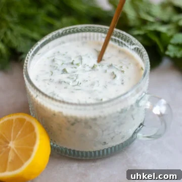 Vegan Creamy Yogurt and Herb Dressing Recipe 8 a glass round bowl of creamy yogurt herb salad dressing including vegan yogurt, lemon juice, maple syrup, mint and dill and salt and pepper sits on a gray surface along with a half of a fresh lemon and some fresh dill and mint bunches in the background