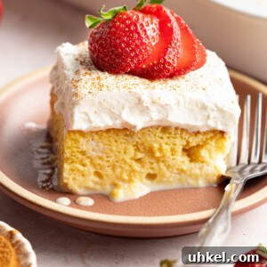 A slice of tres leches cake with a sliced strawberry on top on a plate with a fork for serving.