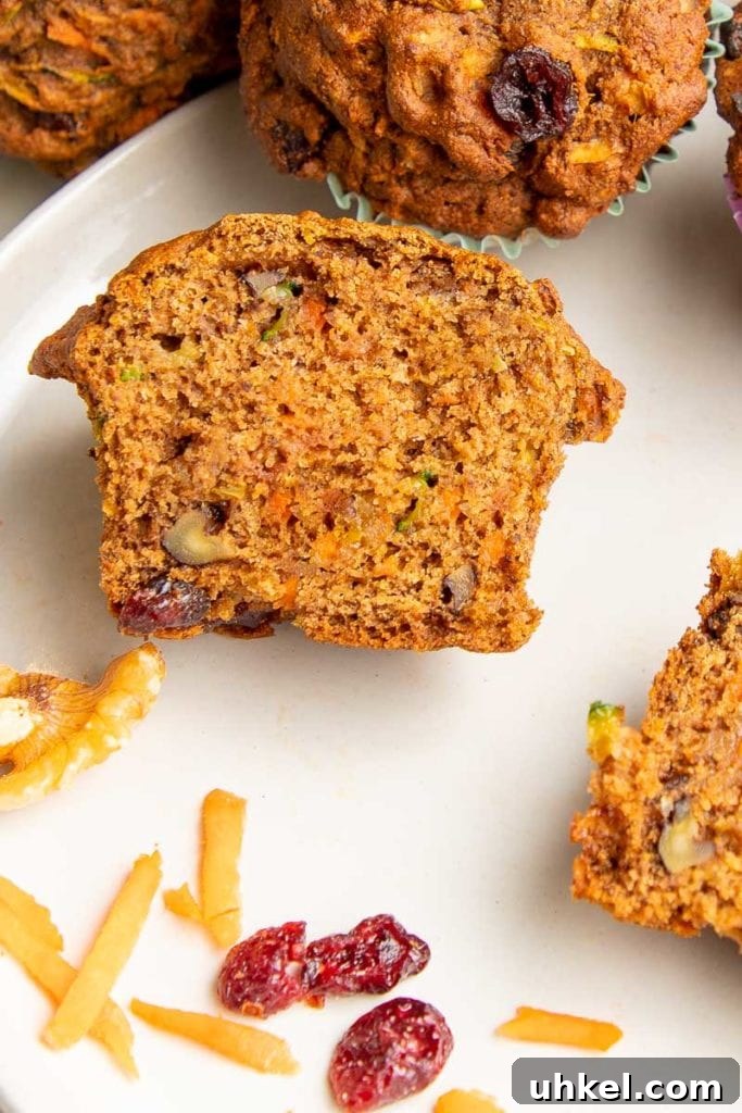 Morning Glory Muffins with Carrots, Apples & Nuts 11 Breakfast muffin that has been cut in half. You can see pieces of shredded carrots and zucchini plus walnuts and dried cranberries. There are a few cranberries and shredded carrots in the foreground on a white plate.