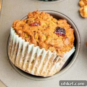 Morning Glory Muffins with Carrots, Apples & Nuts 12 close up of a morning glory muffin, showing a dried cranberry and some shredded carrots. The muffin has a light blue paper liner sitting just outside the pan it was baked in.