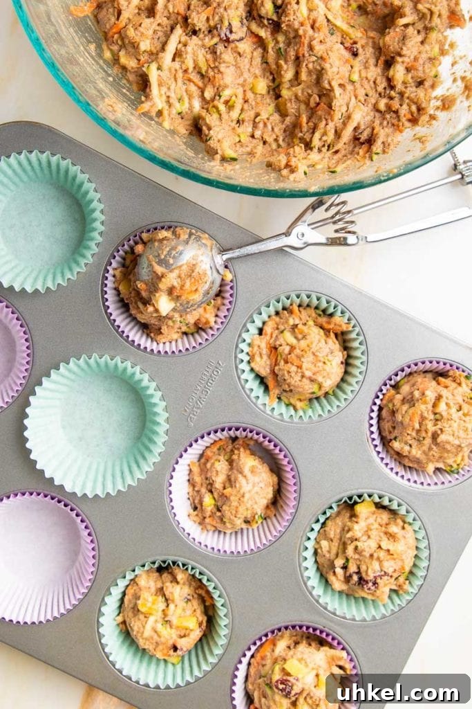 Morning Glory Muffins with Carrots, Apples & Nuts 6 Bowl of muffin batter and a muffin tin lined with purple and light blue liners, half of them already filled with batter and a large cookie scoop filling another.