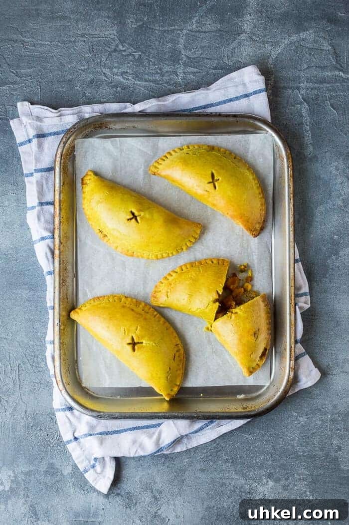 Jamaican vegetable patties - these spiced, Caribbean style vegan vegetable pasties are full of flavour and make a great snack or light meal. Eggless and dairy-free. #veganmeals #veganbaking #meatfree #plantbased Four vegan Jamaican vegetable patties on a baking sheet on a blue and white tea towel.