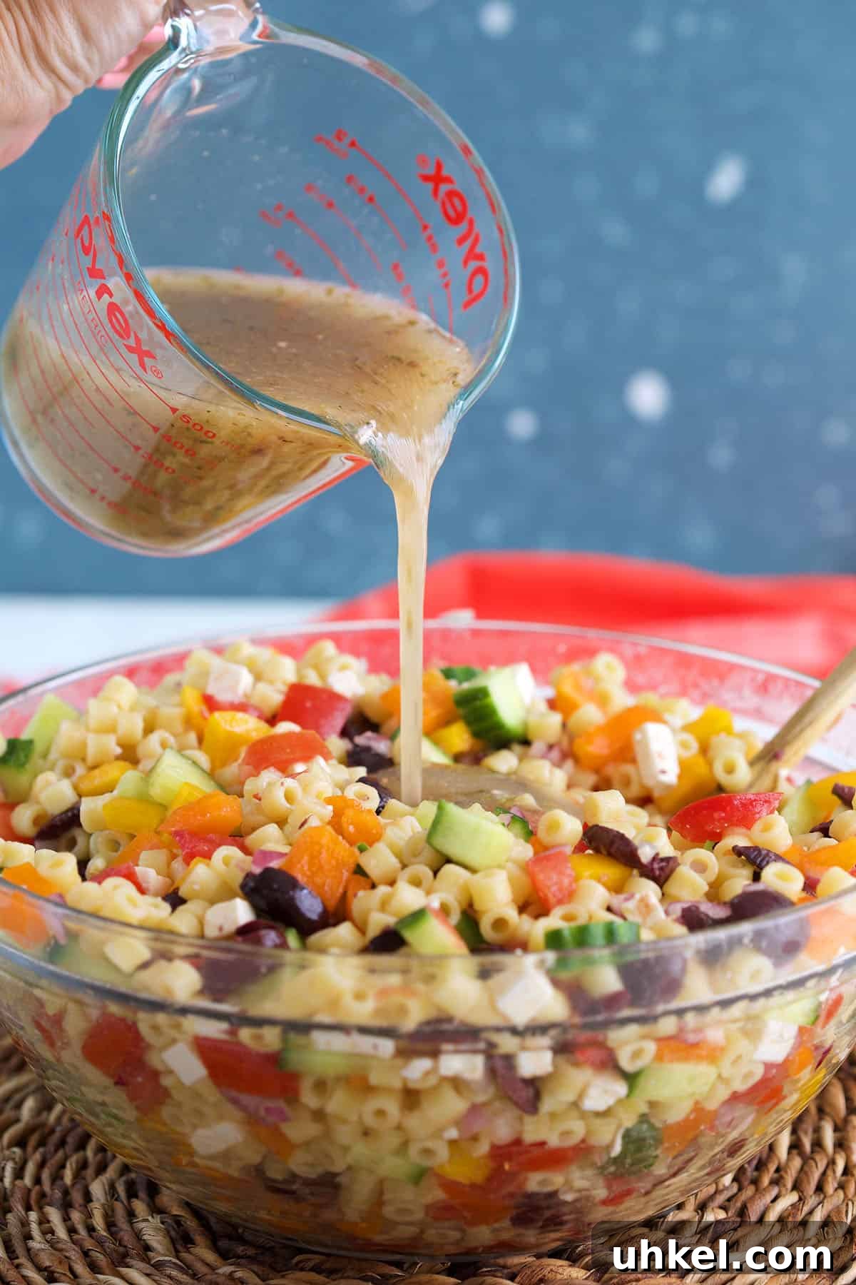 Greek Pasta Salad with Feta, Olives & Fresh Tomatoes 5 Greek Salad Dressing being poured over pasta salad in a glass bowl.