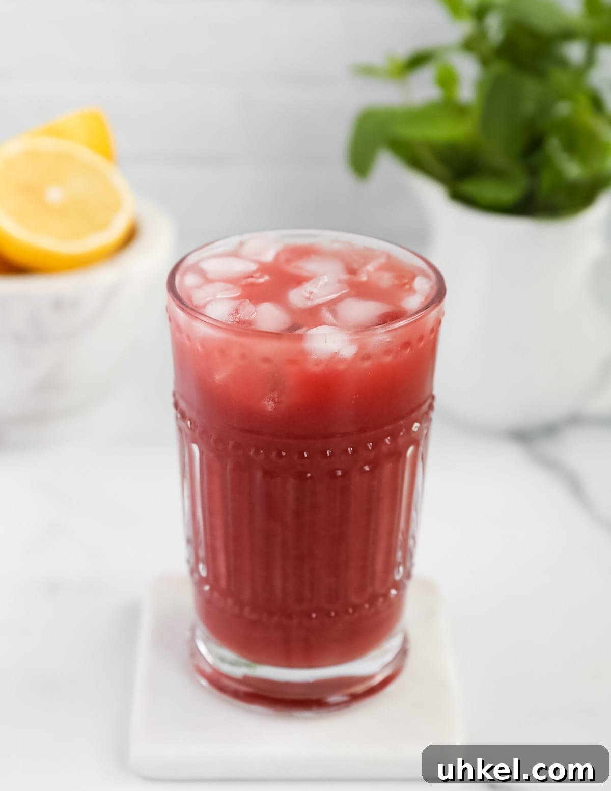 Homemade Strawberry Acai Lemonade 6 A picture of a clear glass filled with ice and pink lemonade. There is fresh mint and lemons in the background.