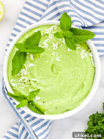 A round white bowl filled with a green kale smoothie. The bowl is sitting on a blue and white stripped cloth, there is a lemon and a bunch of kale in the background.